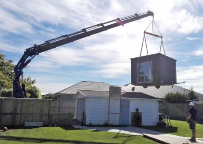 A crane lifting a portable cabin over a house into the back yard