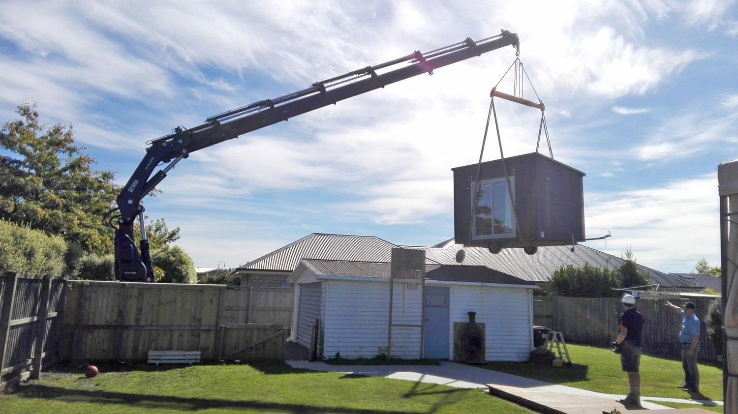 A crane lifting a portable cabin over a house into the back yard